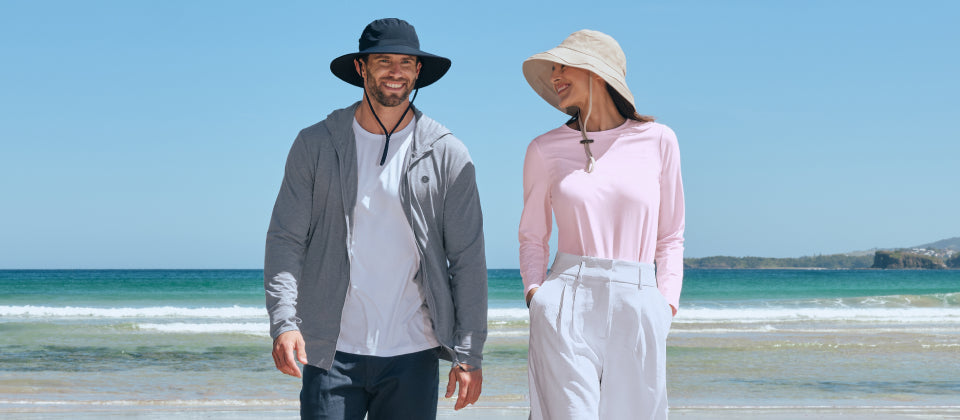 Man and woman walking on a beach wearing Solbari sun hats and UPF 50+ clothing, with clear blue sky and ocean.