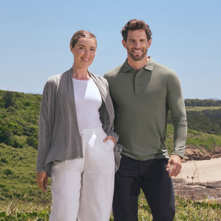 Man and woman wearing Solbari sun protective clothing standing on a grassy area with a scenic beach and mountains in the background.