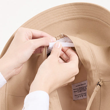 Person adjusting a beige hat with a visible brand label on a white background