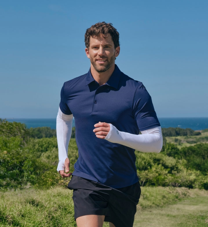 Man running outdoors with a blue polo shirt and black shorts against a scenic background wearing sun protective arm sleeves.