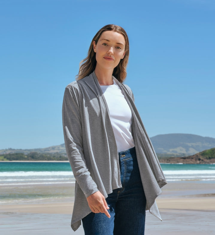 Woman wearing a grey Solbari UPF 50+ sun protective cardigan over a white shirt with a beach and blue sky in the background.