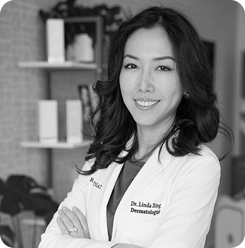 Black and white photo of a woman in a white lab coat with 'Dr. Linda Xing' on it, standing in an office setting.