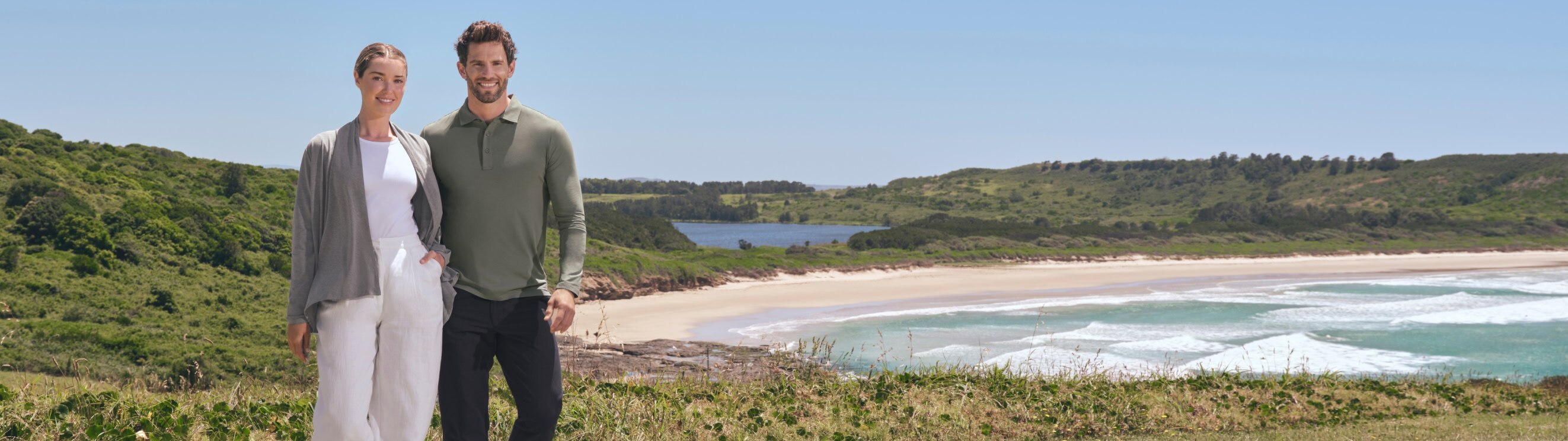 Man and woman wearing Solbari sun protective clothing standing on a grassy area with a scenic beach and mountains in the background.